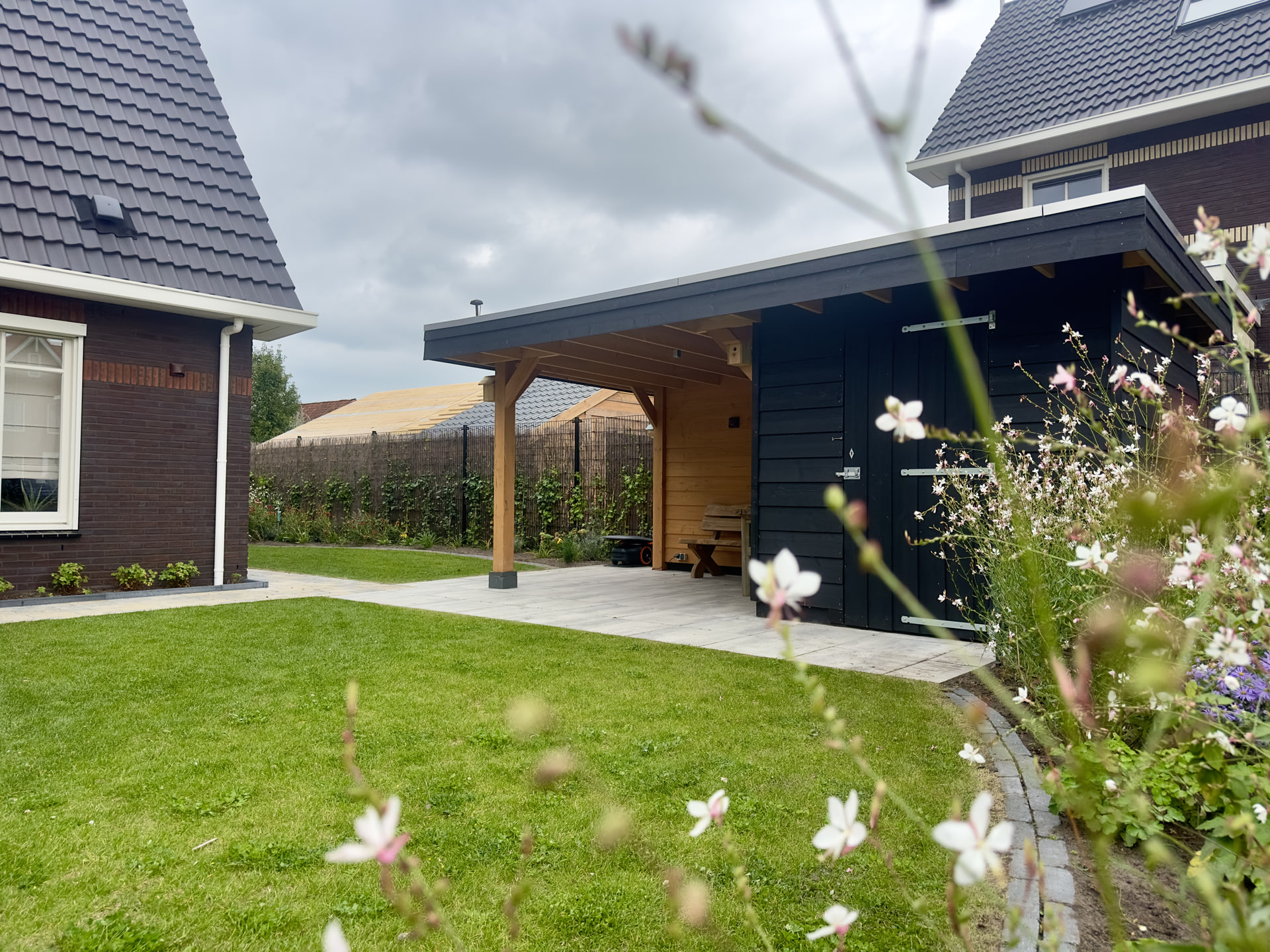 Achtertuin in Gorredijk met gazon, een terras van lichte tegels onder een houten overkapping met zwarte berging en een border met witte gaura op de voorgrond